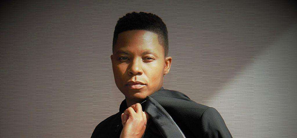 Milon V Parker dressed in black, standing against a grey textured wall
