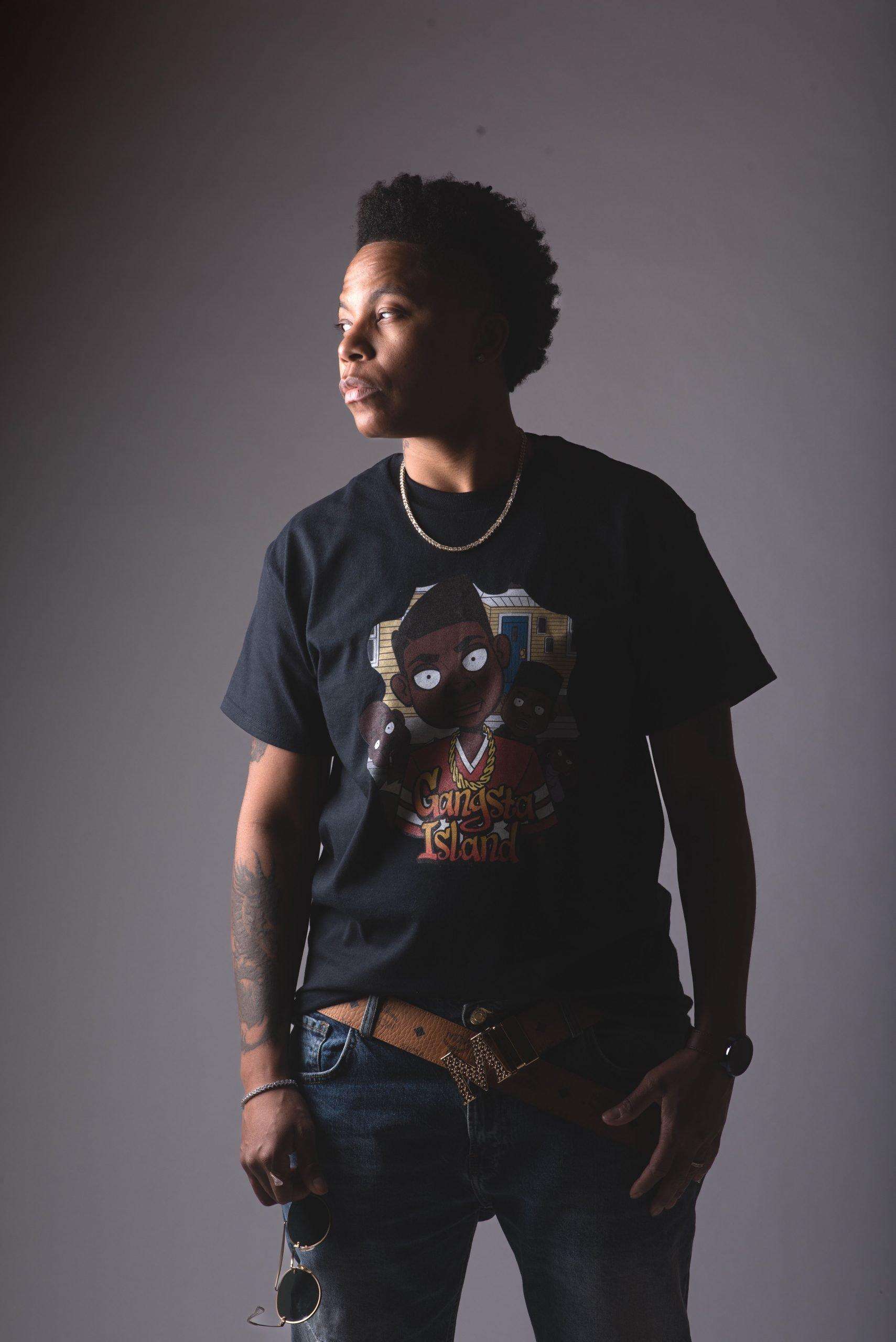 Milon V Parker modeling in a black Gangsta Island t-shirt, posing in front of a grey wall